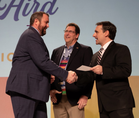 John Aird, in a charcoal suit, shakes the hand of a man who gives him an award
